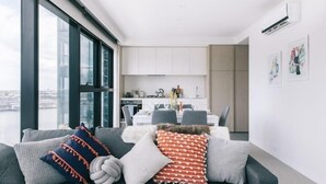 Een moderne woonkamer met een grijze bank, kussens met een wit en rood patroon, een eethoek met een witte tafel en stoelen, en een stadszicht door grote ramen.