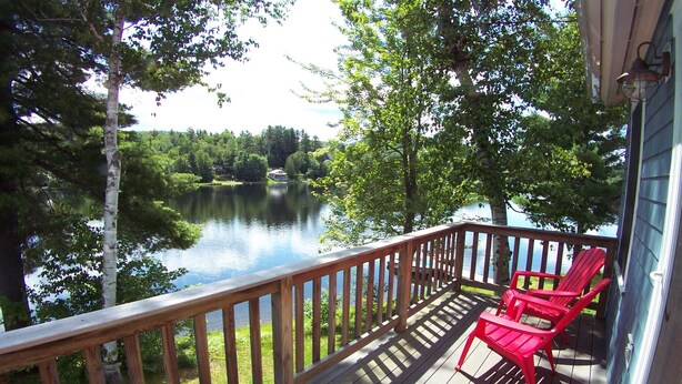 Une terrasse en bois dotée d’une balustrade donnant sur un lac, avec des fauteuils Adirondack rouges.