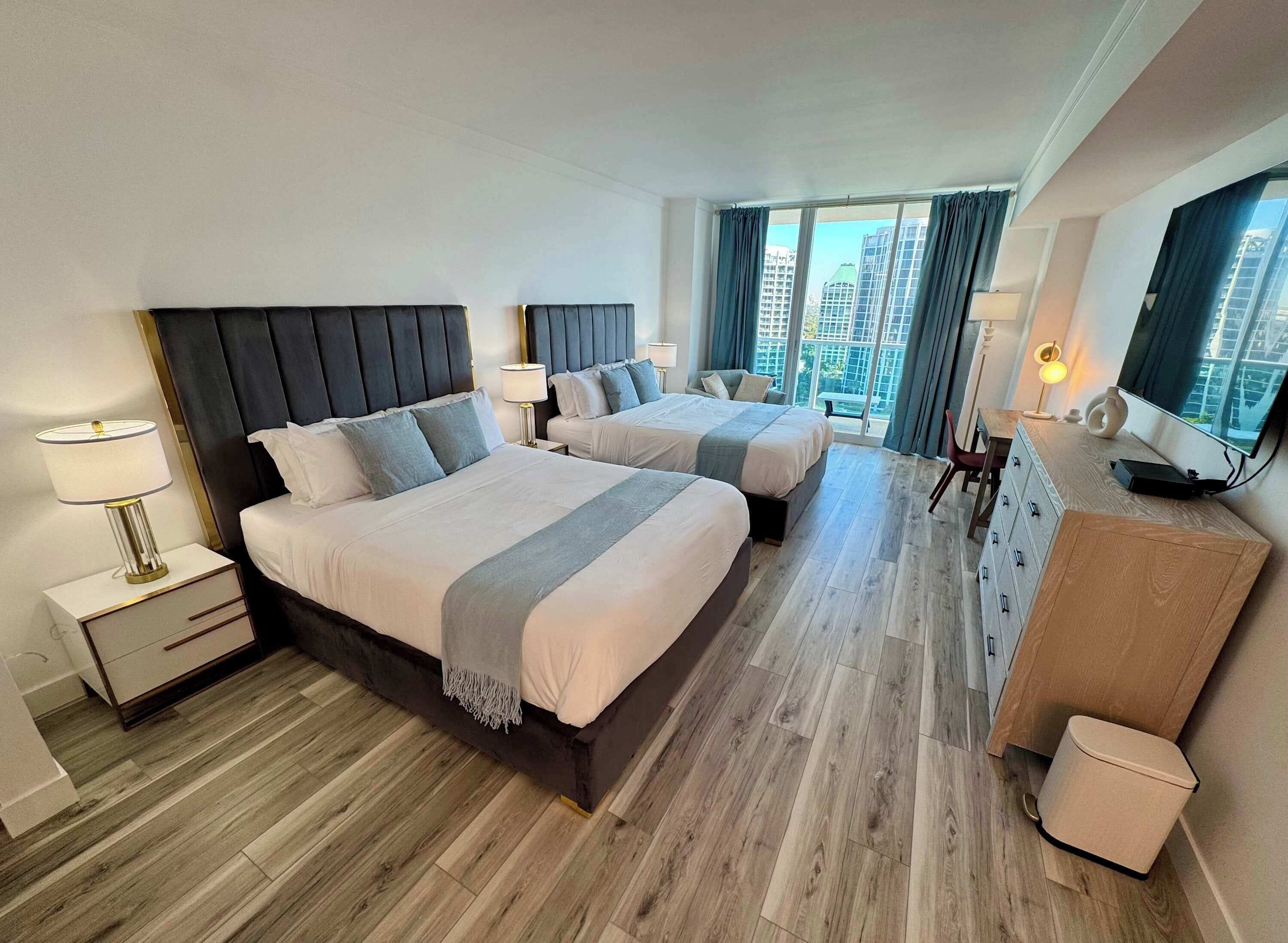 A hotel room with two beds, a TV, a desk, and a view of the city.