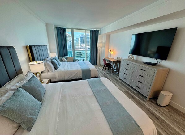 Habitación de hotel con dos camas, un escritorio, televisión y vista a la ciudad.