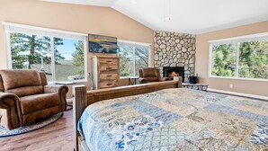 A spacious bedroom with a large bed, a stone fireplace, a television, and a view of trees through large windows. - Moonridge Memories - 5 Bedroom, Hot Tub, Game Room, Pool Table (Big Bear City)