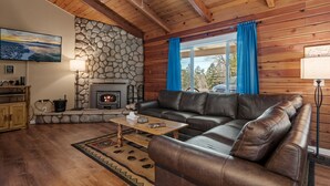 A cozy living room with a stone fireplace, leather sofa, and a large window with blue curtains. - Moonridge Memories - 5 Bedroom, Hot Tub, Game Room, Pool Table (Big Bear City)