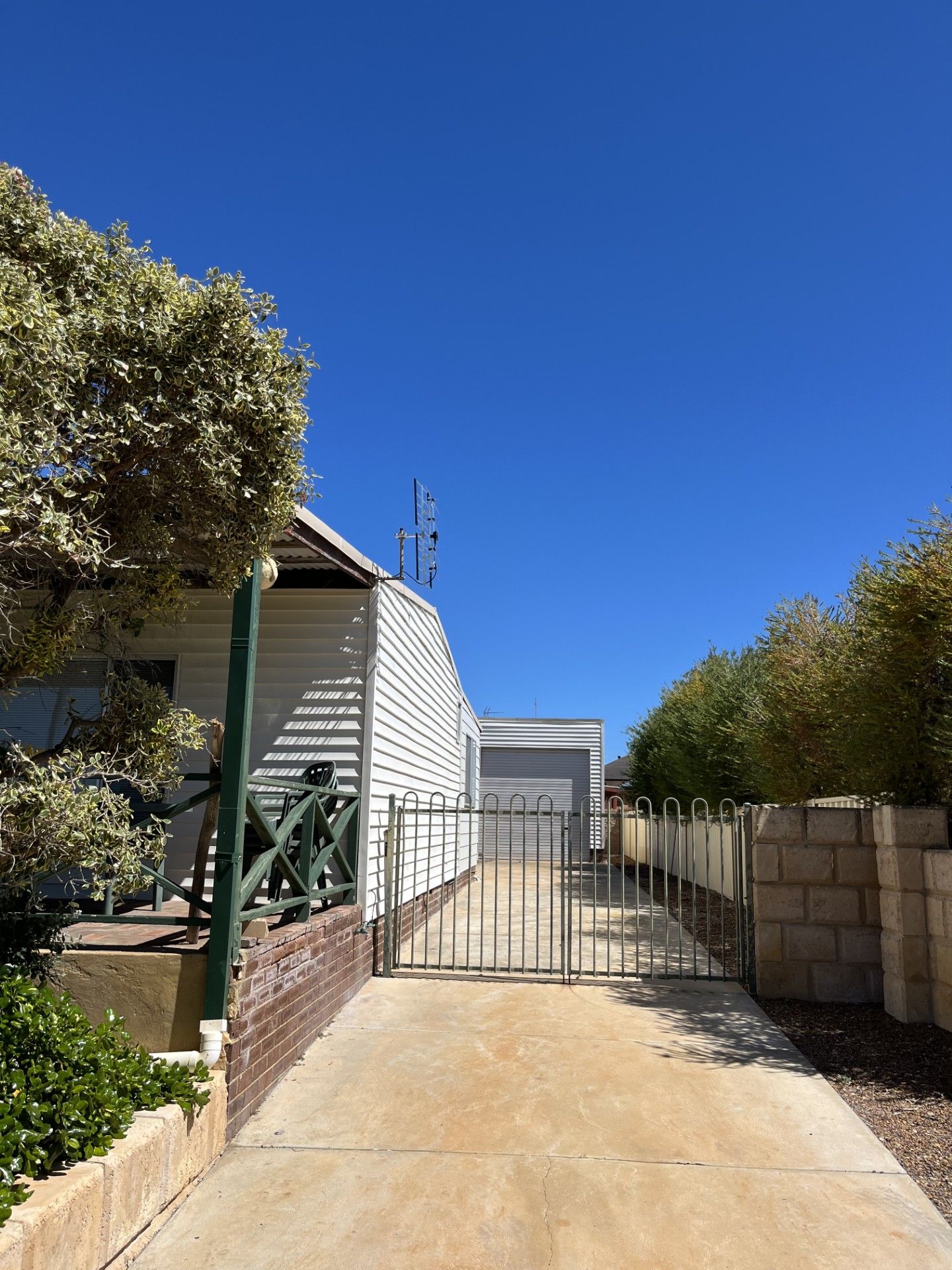 Une allée fermée par un portail mène à une propriété résidentielle dotée d’un garage, le tout étant entouré de verdure et surplombé par un ciel bleu dégagé.
