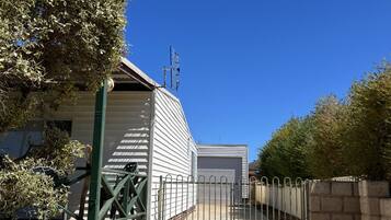 Une allée fermée par un portail mène à une propriété résidentielle dotée d’un garage, le tout étant entouré de verdure et surplombé par un ciel bleu dégagé.