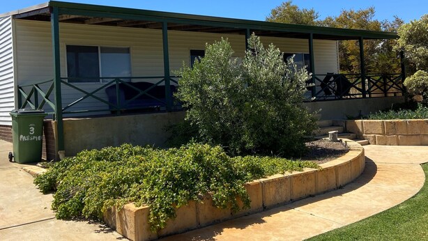 A single-story building with a covered patio area, a small garden with a bush, and a concrete pathway leading to the entrance.
