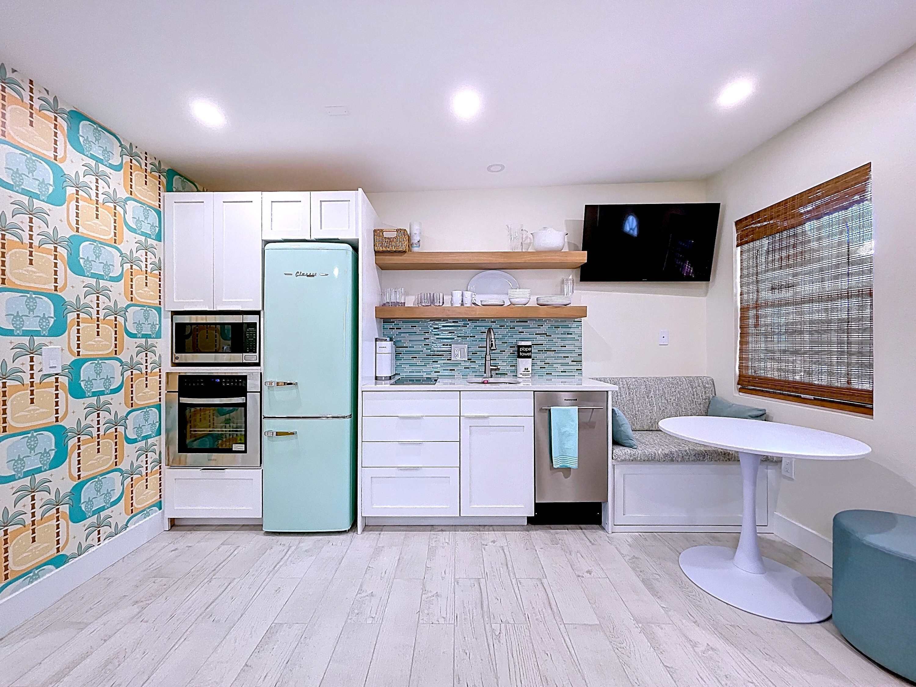 A compact kitchen with white cabinets, a teal refrigerator, a microwave, and a built-in oven. There's a small dining area with a white table and a single chair.