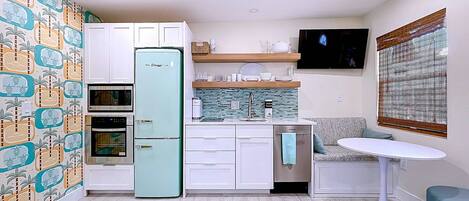 A compact kitchen with white cabinets, a teal refrigerator, a microwave, and a built-in oven. There's a small dining area with a white table and a single chair.