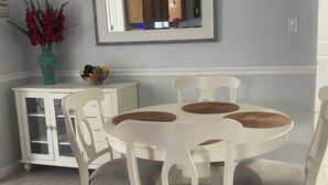 Un comedor con una mesa y sillas blancas, un jarrón con flores rojas y un frutero sobre un mueble blanco.