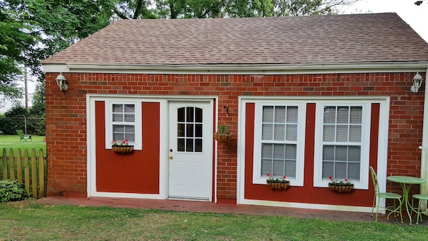 Un petit bâtiment en briques, à l’extérieur rouge, avec une porte blanche et des fenêtres dotées de volets. Deux jardinières suspendues ornent les rebords des fenêtres.