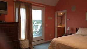 Un dormitorio con una cama grande, un aparador, un ventanal con cortinas, un baño con lavamanos y espejo, y vistas a una terraza y una casa.