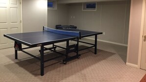 Una mesa de ping pong en una habitación con piso alfombrado, una puerta y un ventanal con persianas.