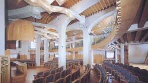 Un salón amplio con filas de sillas y un techo alto con vigas de madera y lámparas colgantes.