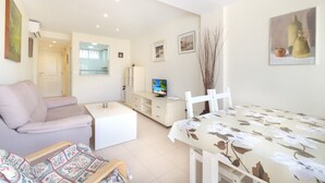 A modern living room with a sofa, a dining table with a floral tablecloth, a TV on a stand, and a painting on the wall.