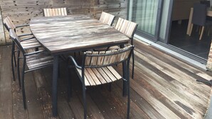 Terraza de madera exterior con una mesa grande y sillas, puerta de cristal que da a un espacio interior y pared de ladrillo.
