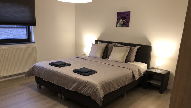Una cama bien tendida con cabecera, dos mesitas de noche y una ventana con persianas.