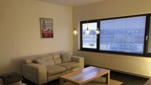 Una sala de estar con un sofá, una mesa de centro y un radiador. Hay una ventana con persianas y un cartel en la pared.