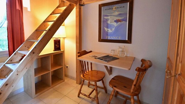 Una escalera de madera conduce a una habitación con una mesa, dos sillas, un cartel enmarcado y una lámpara.