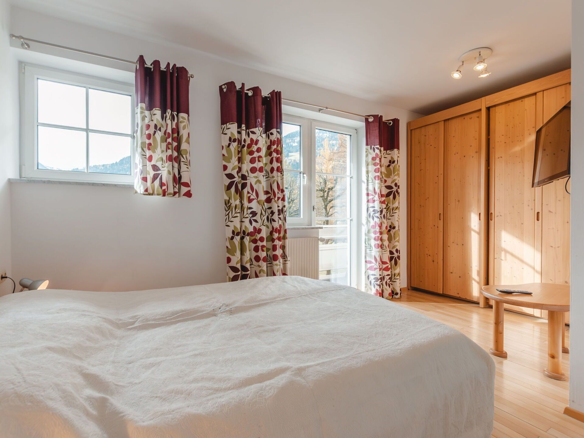A bedroom with a large bed, two windows with curtains, a wooden wardrobe, and a TV mounted on the wall.