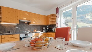 Une cuisine moderne avec des meubles en bois, une table à manger agrémentée d’un panier de fruits, et une vue sur un paysage montagneux grâce à de grandes fenêtres.