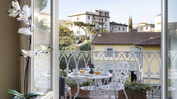 Un balcon avec des chaises et des tables en métal peintes en blanc, des plantes en pot et une vue sur les bâtiments qui se dressent à l’arrière-plan.