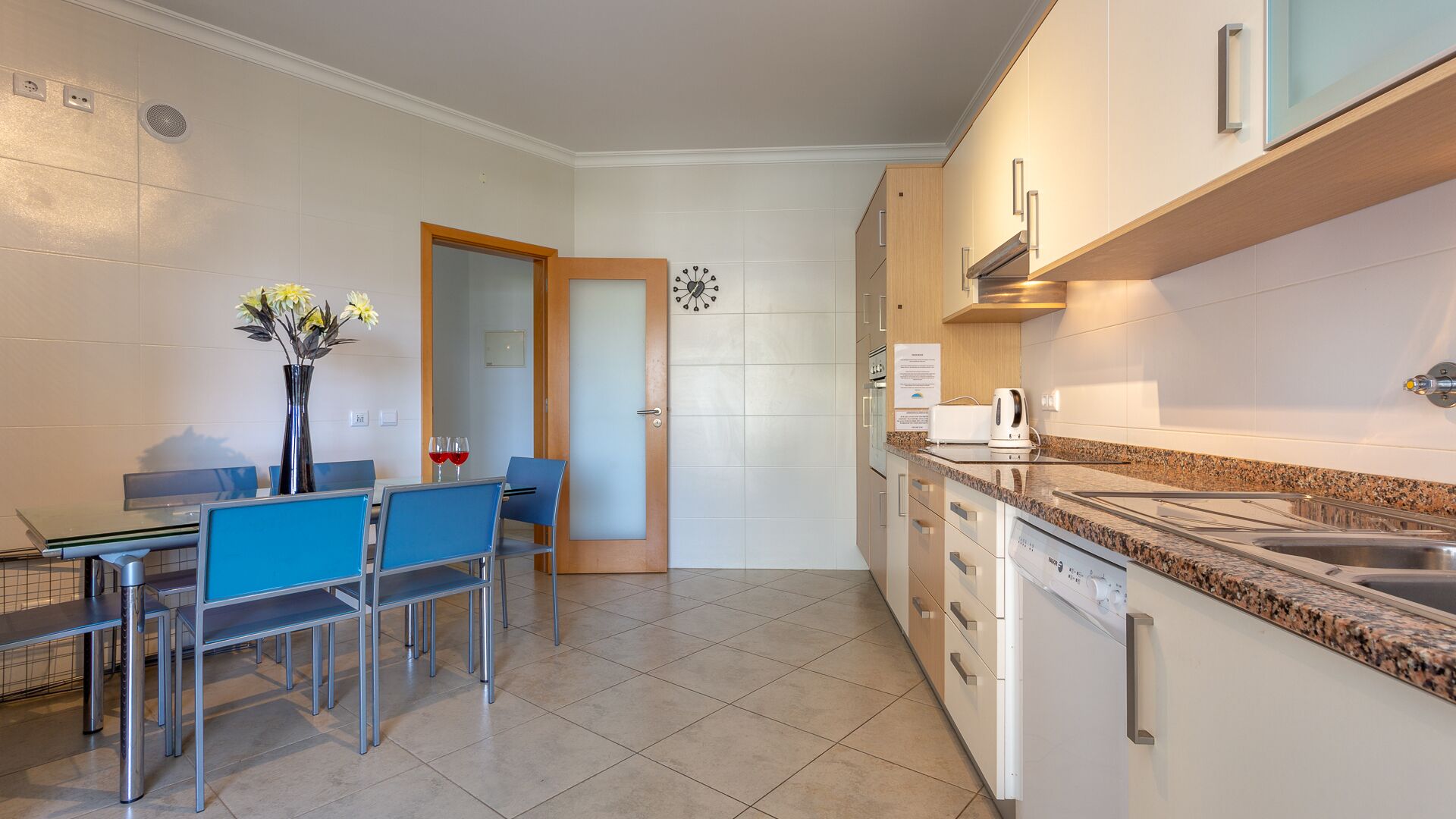 A modern kitchen with a granite countertop, stainless steel appliances, and a dining area with blue chairs and a glass table.