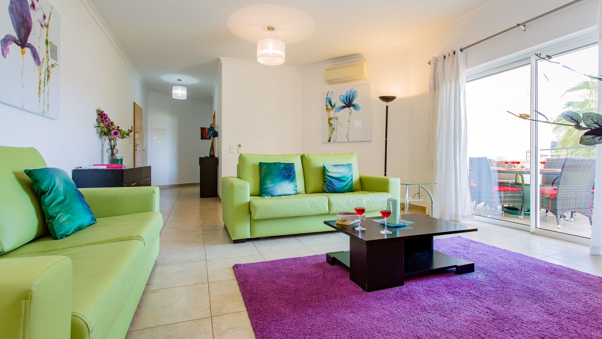 A modern living room with a green sofa set, a coffee table, and a purple rug. There are paintings on the walls and a view of the outdoors through the glass door.