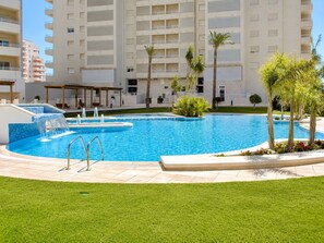 A swimming pool with a water feature, surrounded by a grassy area and palm trees, with a multi-story building in the background. - Calpe: apartment/ flat - Calpe (Calpe)