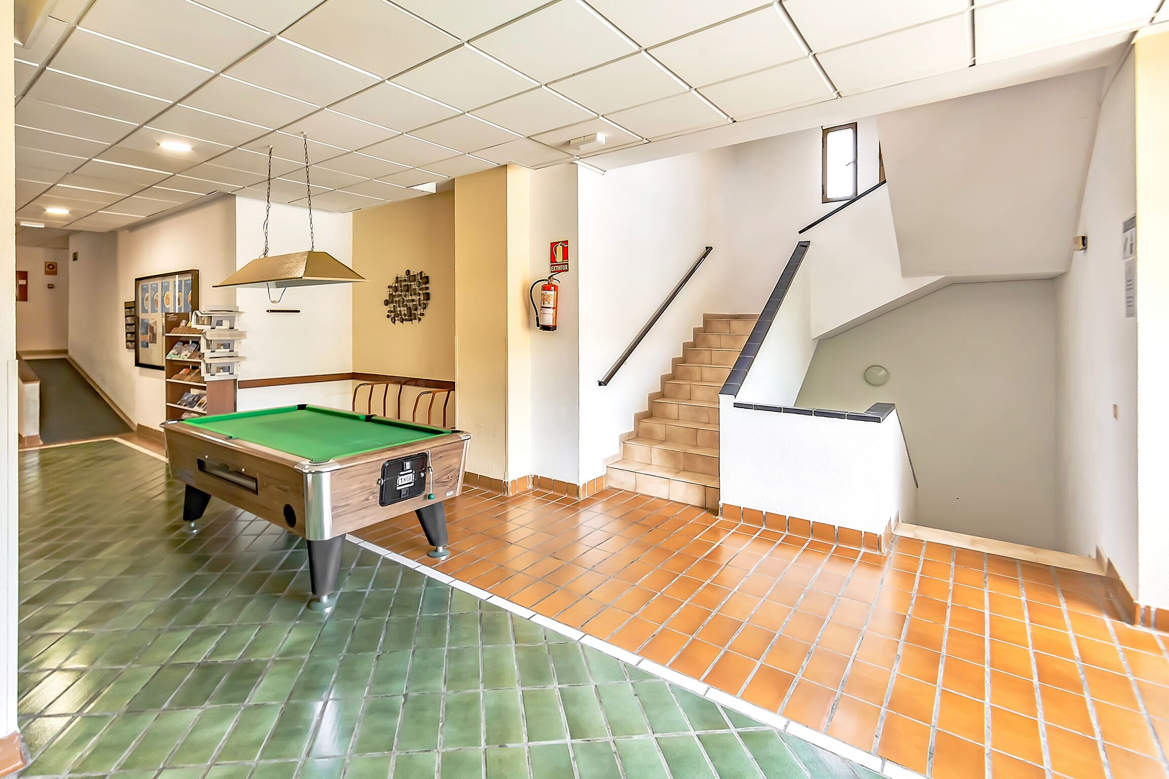 A pool table in a well-lit room with a tiled floor, a staircase, and a fire extinguisher on the wall.