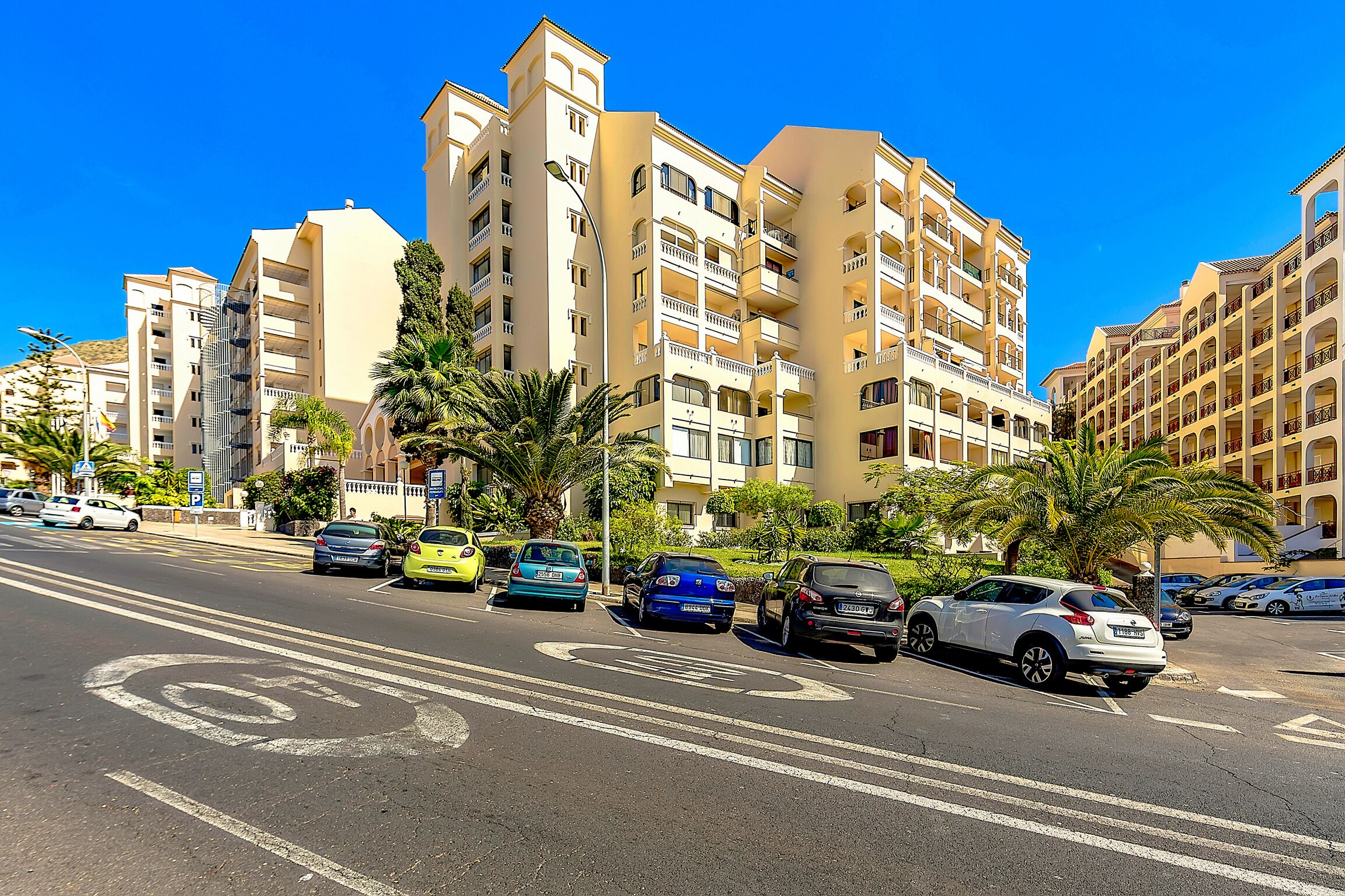 A multi-story hotel with balconies, parked cars, and palm trees.