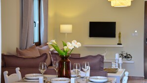 A dining area with a table set for four, a vase with flowers, a sofa, a television, and a bicycle in the background. - Elianthos Villas / Villa Rose 🌹 (Chania)