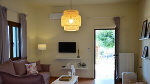 A living room with a sofa, a coffee table, a TV, a hanging light fixture, and a view of the outdoors. - Elianthos Villas / Villa Rose 🌹 (Chania)