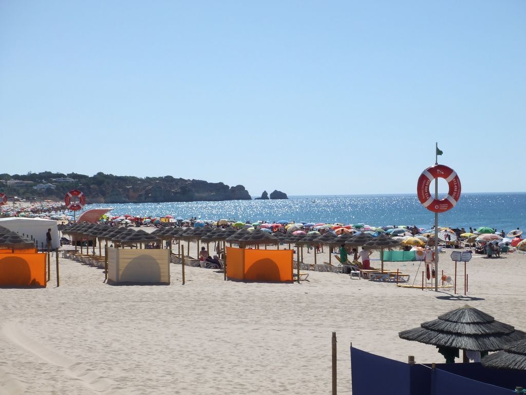 Une plage dotée de nombreux parasols et de cabanes de plage, d’un panneau indiquant la présence d’une bouée de sauvetage, et offrant une vue dégagée sur la mer.