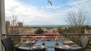 Una mesa de comedor dispuesta con platos, vasos y cubiertos en un balcón con vistas a un paisaje urbano y al mar.
