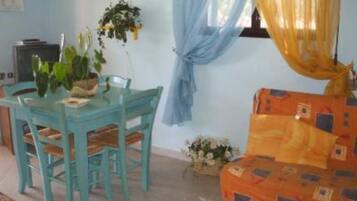 Ein Zimmer mit einem blaugrünen Esstisch und Stühlen, einem kleinen orangefarbenen Sofa und einem Fenster mit blauen Vorhängen.