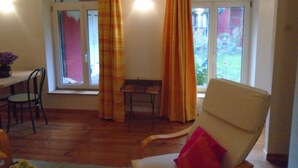 A room with a wooden floor, a white chair with a red pillow, a dining table with chairs, and a window with curtains. - To explore the Christmas Markets in Alsace! (Neuf-Brisach)