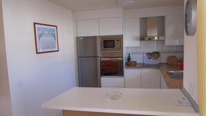 Cocina compacta con gabinetes blancos, un refrigerador de acero inoxidable, un horno y un microondas. Hay una pequeña isla con taburetes, un cuadro en la pared y un reloj.