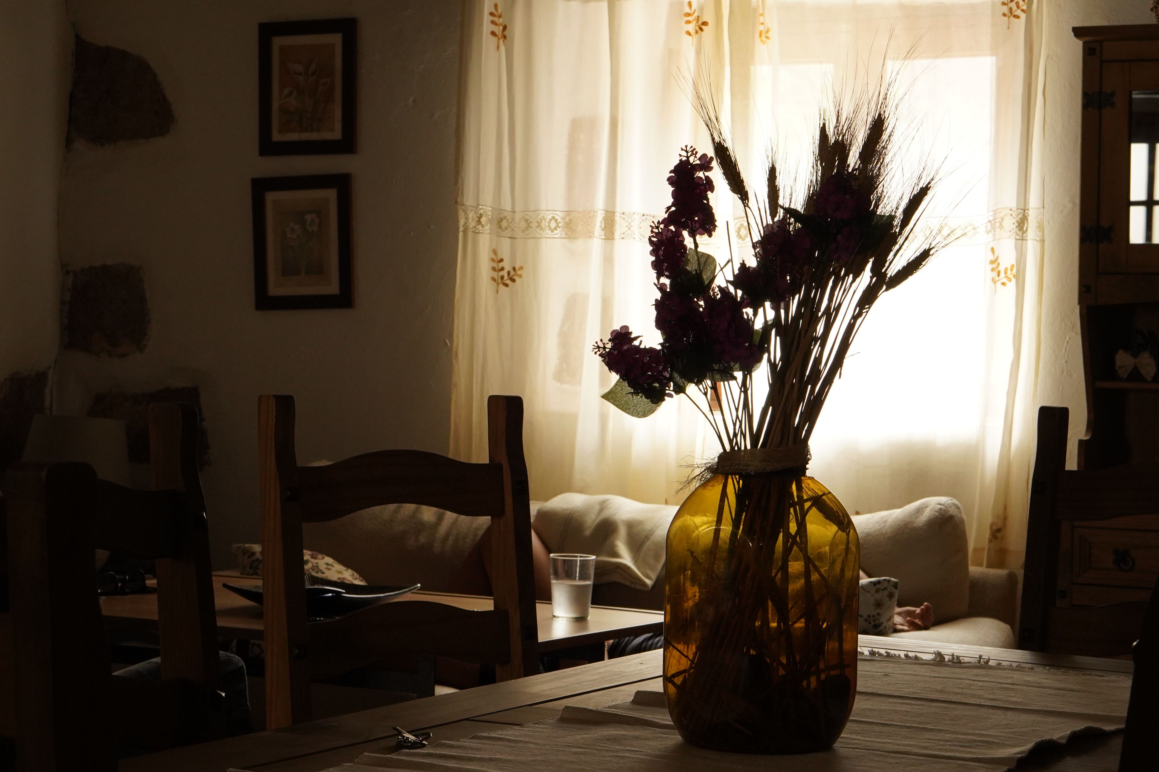 Una habitación con una mesa de madera, un florero con plantas secas, un sofá y una ventana con cortinas.