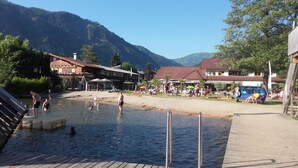 Una terraza de madera que da a una zona para nadar, con gente en el agua, rodeada de edificios y montañas.