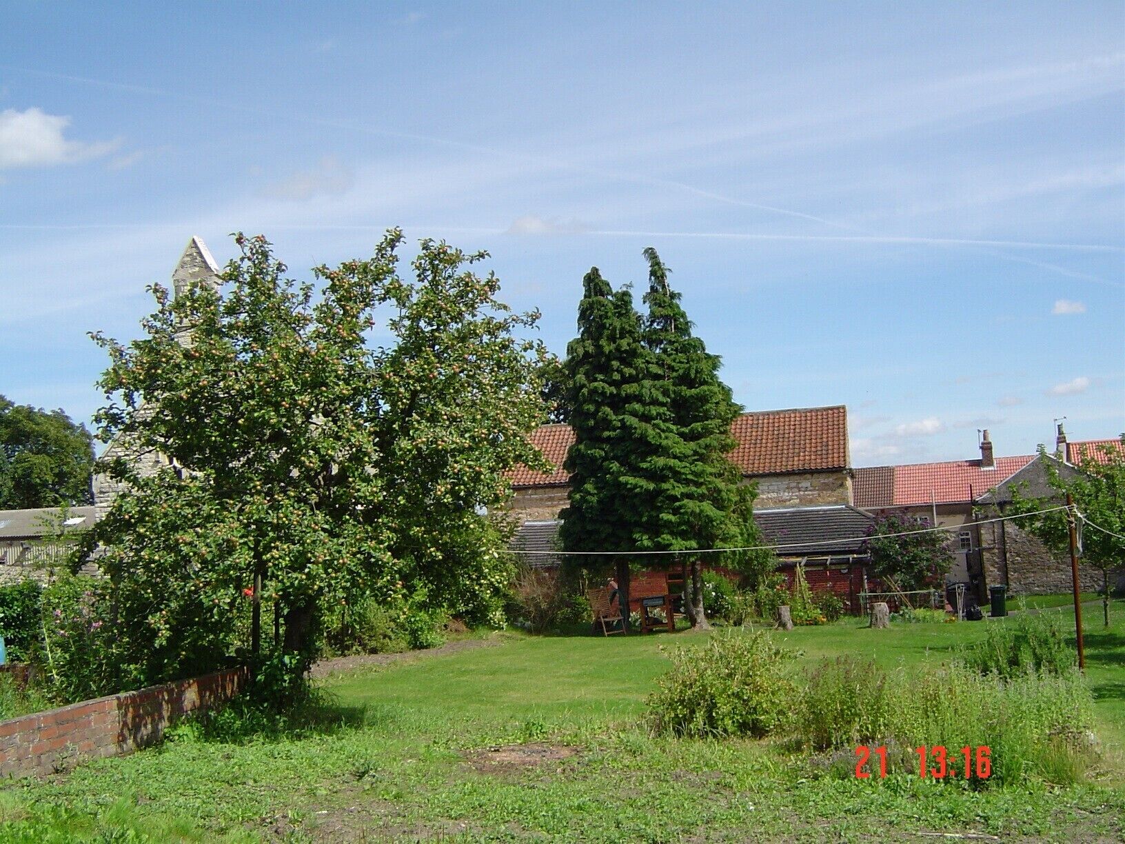 Un jardin avec une pelouse, un grand arbre à feuilles persistantes et un bâtiment en pierre surmonté d’un toit recouvert de tuiles rouges.