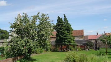 Un jardin avec une pelouse, un grand arbre à feuilles persistantes et un bâtiment en pierre surmonté d’un toit recouvert de tuiles rouges.