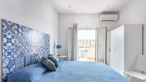 Un dormitorio con un cabecero estampado, una cama con ropa de cama azul, una mesita de noche y vistas al exterior.