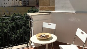 Un balcón con una mesa redonda y dos sillas blancas, con vista a un paisaje urbano que incluye edificios y montañas.