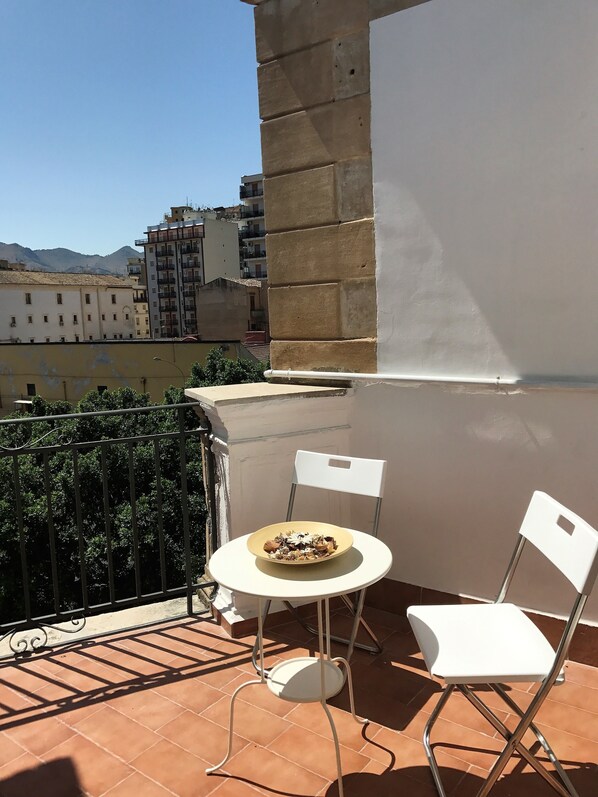 A balcony with a round table and two white chairs, overlooking a cityscape with buildings and mountains.