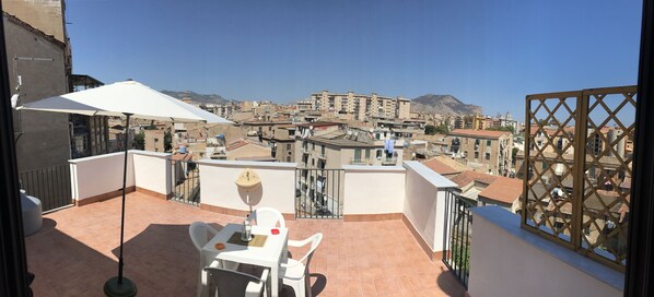 A terrace with a table and chairs, a white umbrella, and a view of a cityscape with buildings and mountains. - Frank's house - flat with terrace in the centre (Palermo)