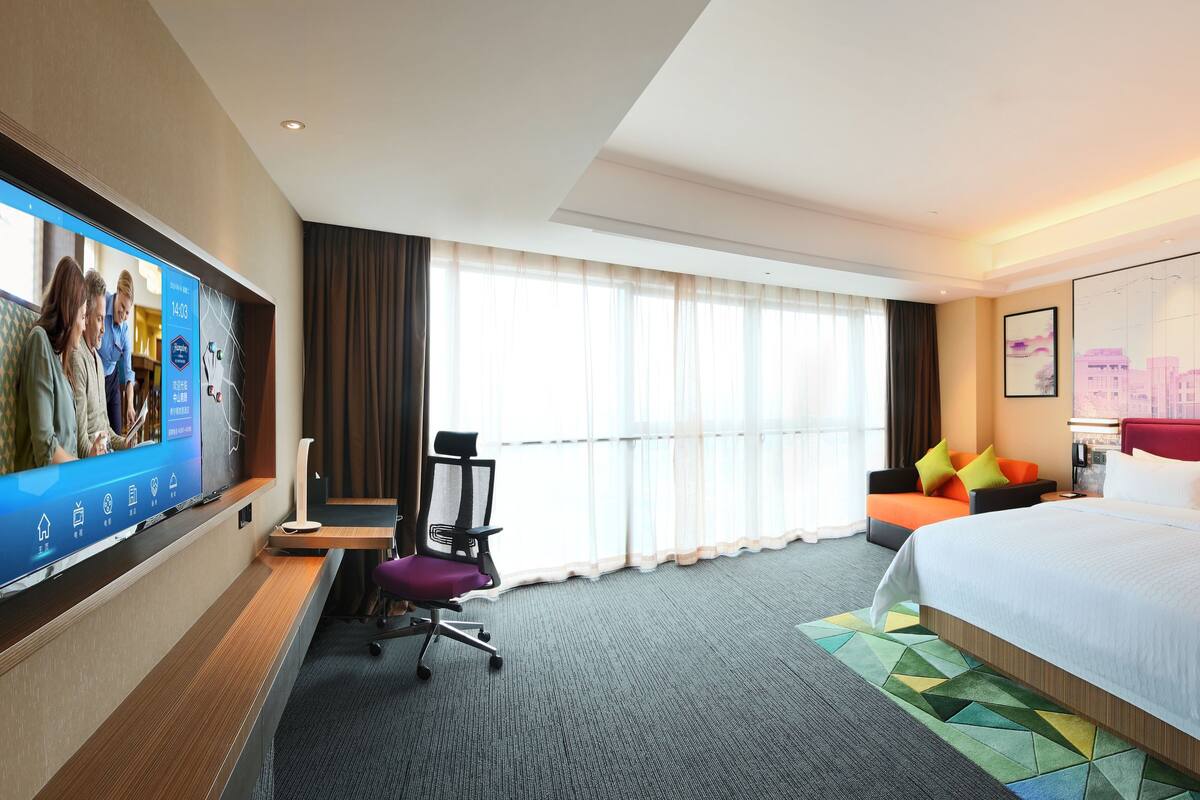 Habitación de hotel con un televisor de pantalla plana grande, una cama con sábanas blancas, un escritorio con silla y un ventanal con cortinas.