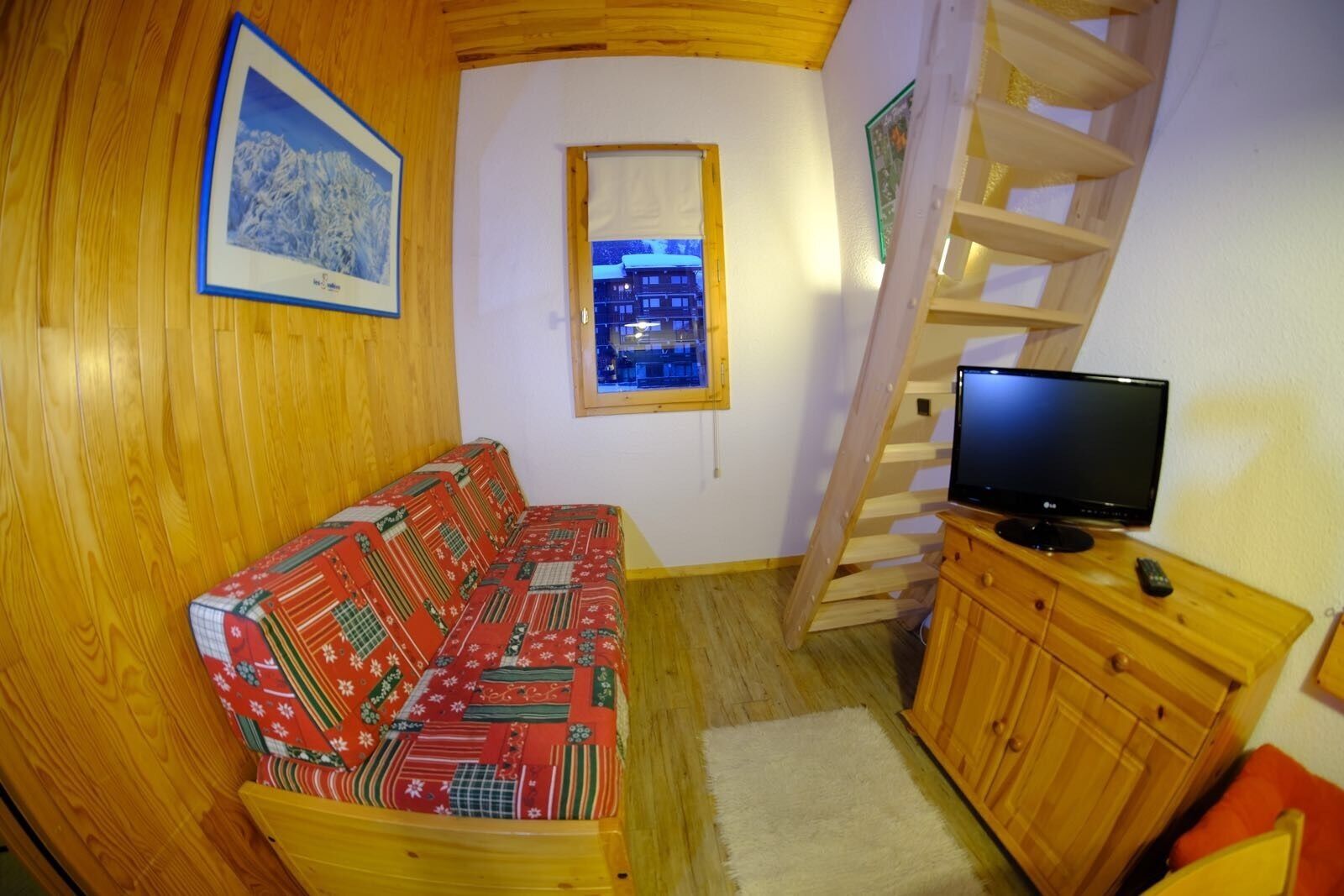 Ein gemütliches Zimmer mit Holzboden, einer gemusterten Couch, einem Fernseher auf einer Holzkommode und einem Fenster mit Blick auf Gebäude.