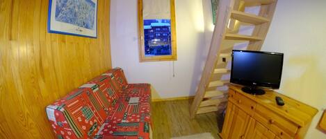 Ein gemütliches Zimmer mit Holzboden, einer gemusterten Couch, einem Fernseher auf einer Holzkommode und einem Fenster mit Blick auf Gebäude.