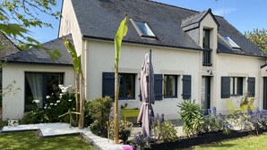 A two-story house with a sloped roof, multiple windows with shutters, and a garden with various plants and a bench. - 4 BEDROOM HOUSE FOR 2 to 6 PEOPLE (La Forêt-Fouesnant)