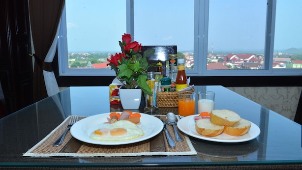 Una mesa de desayuno con platos de huevos, tostadas y un vaso de jugo de naranja, frente a una ventana con vistas a edificios y follaje.