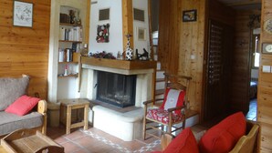 Un salon chaleureux avec une cheminée, des poutres en bois, une bibliothèque et un fauteuil rouge.